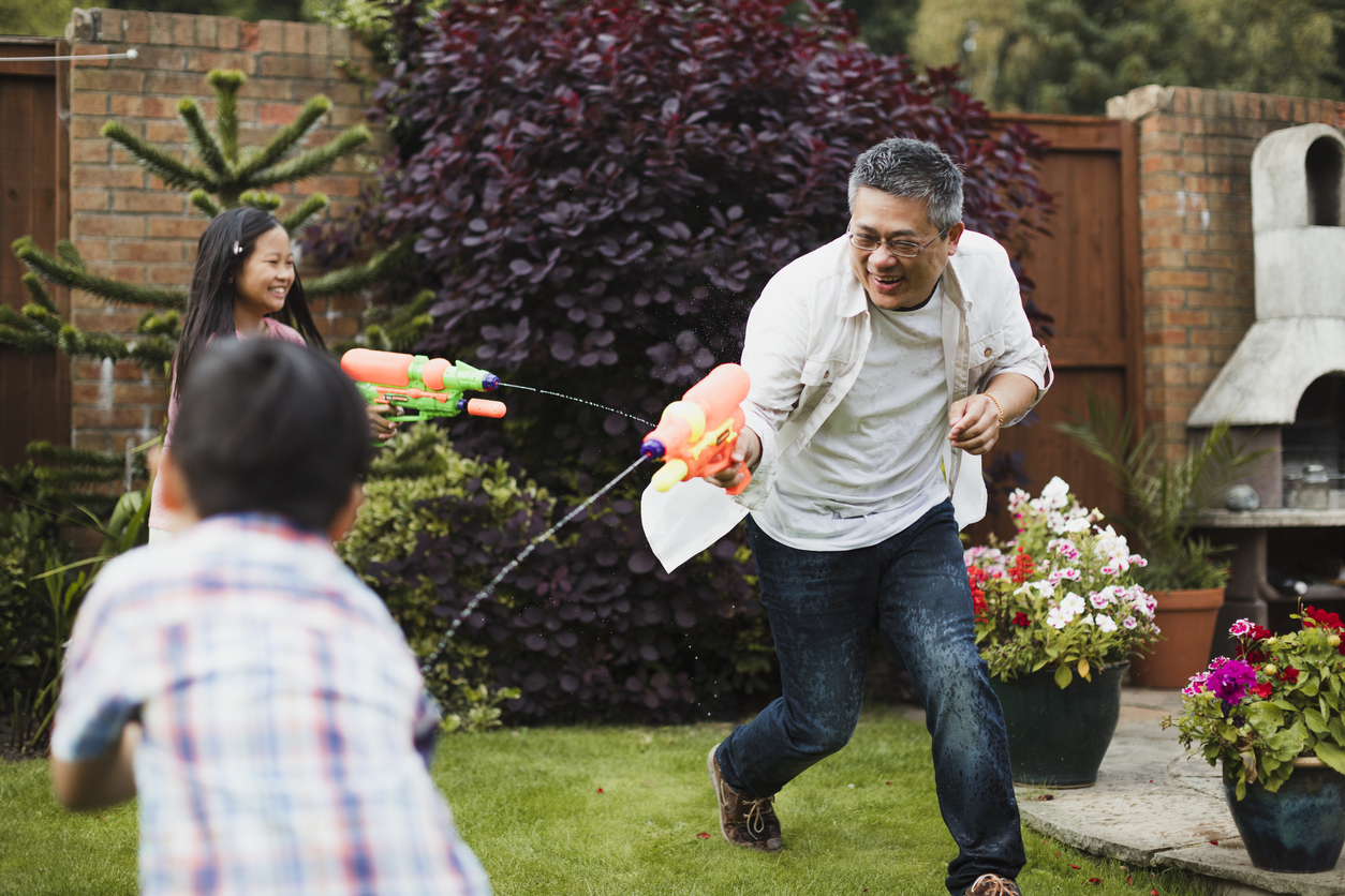 Family are having a water fight together with water pistols in the garden. The little girl is aiming for her dad, who is aiming for the little boy.